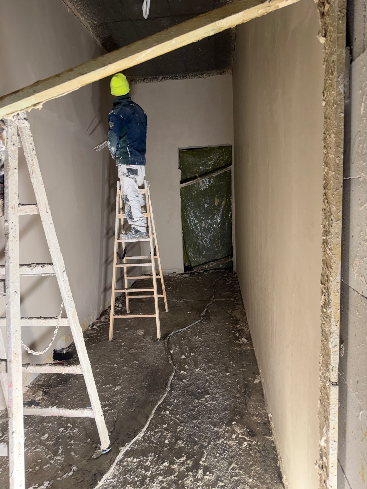Pracownik w żółtej czapce na drabinie, tynkuje ścianę wewnątrz budynku. Widoczne świeżo otynkowane ściany i surowy stan podłogi. Prace wykończeniowe w trakcie.