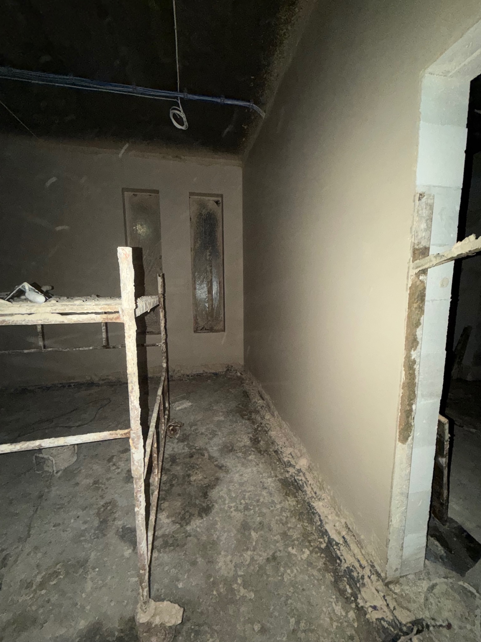 Wnętrze po tynkowaniu maszynowym: gładkie ściany, widoczne ślady prac na podłodze, rusztowanie w rogu. Dwa okienka w ścianie.