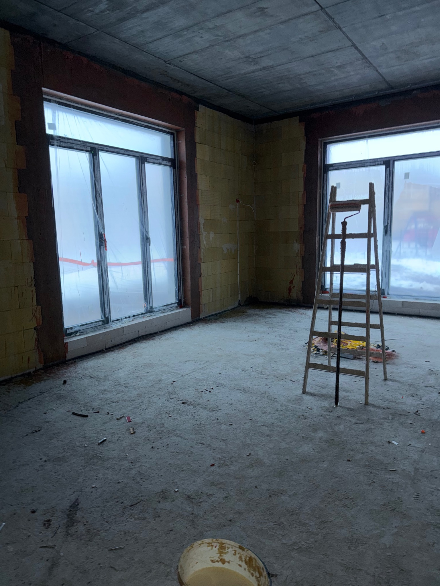 Surowe wnętrze z oknami zabezpieczonymi folią, drabina malarska i wiadro z farbą. Przygotowania do tynkowania w nowym budynku.