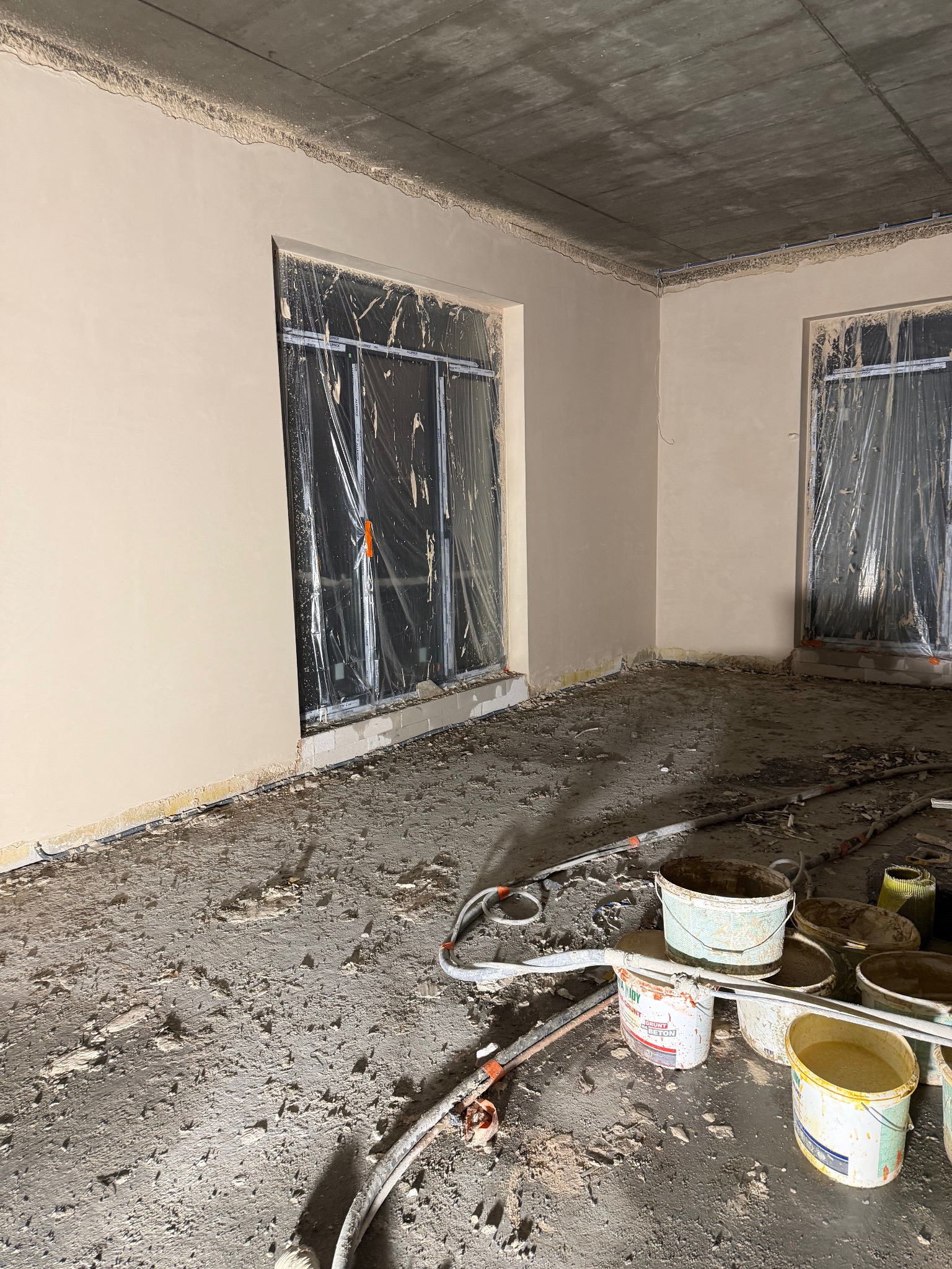 Wnętrze w trakcie prac tynkarskich: ściany pokryte świeżym, jasnym tynkiem, okna zabezpieczone folią, na podłodze widoczne narzędzia i resztki materiałów budowlanych.