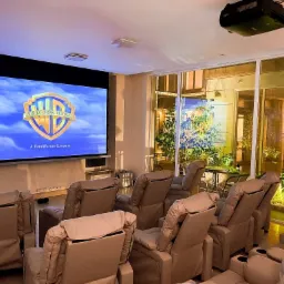 Prywatna sala kinowa z beżowymi fotelami z regulacją, dużym ekranem wyświetlającym logo Warner Bros i widokiem na patio przez szklane ściany.