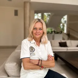 Kobieta z blond włosami siedzi na kanapie z założonymi rękami, ubrana w koszulkę polo z logo 'ZEUS BOX & REHAB' w jasnym, przestronnym wnętrzu.