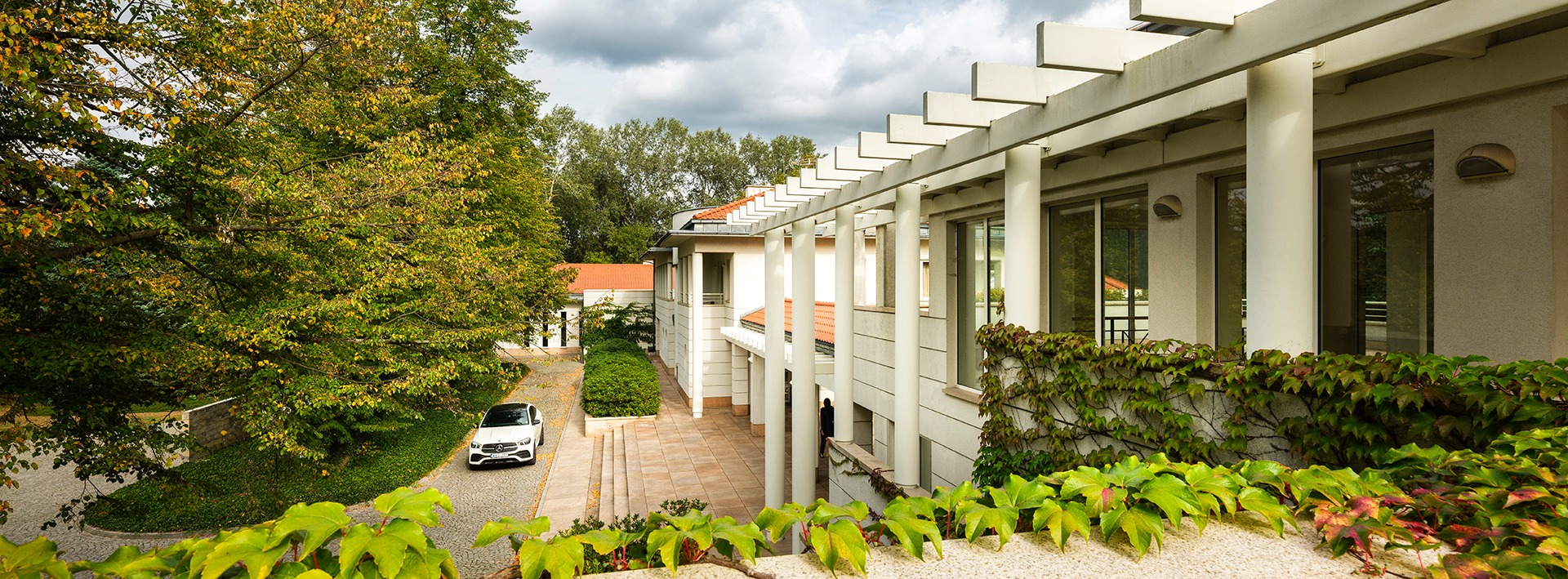 Nowoczesny budynek kliniki z białymi kolumnami i pergolą, otoczony zielenią i drzewami, z białym samochodem zaparkowanym na podjeździe.