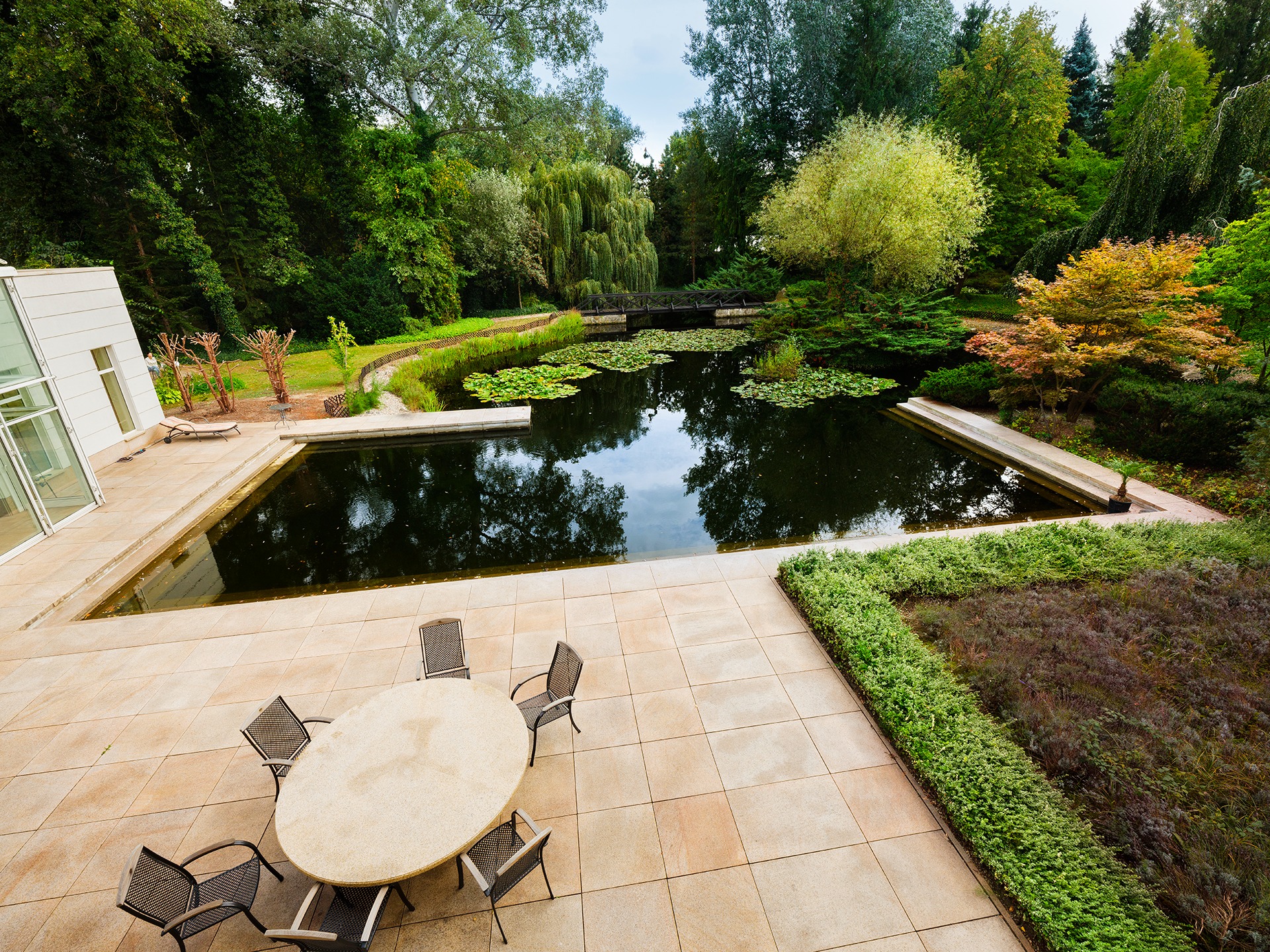 Widok z góry na taras z okrągłym stołem i krzesłami, przylegający do basenu z liliami wodnymi i bujną zielenią w tle.