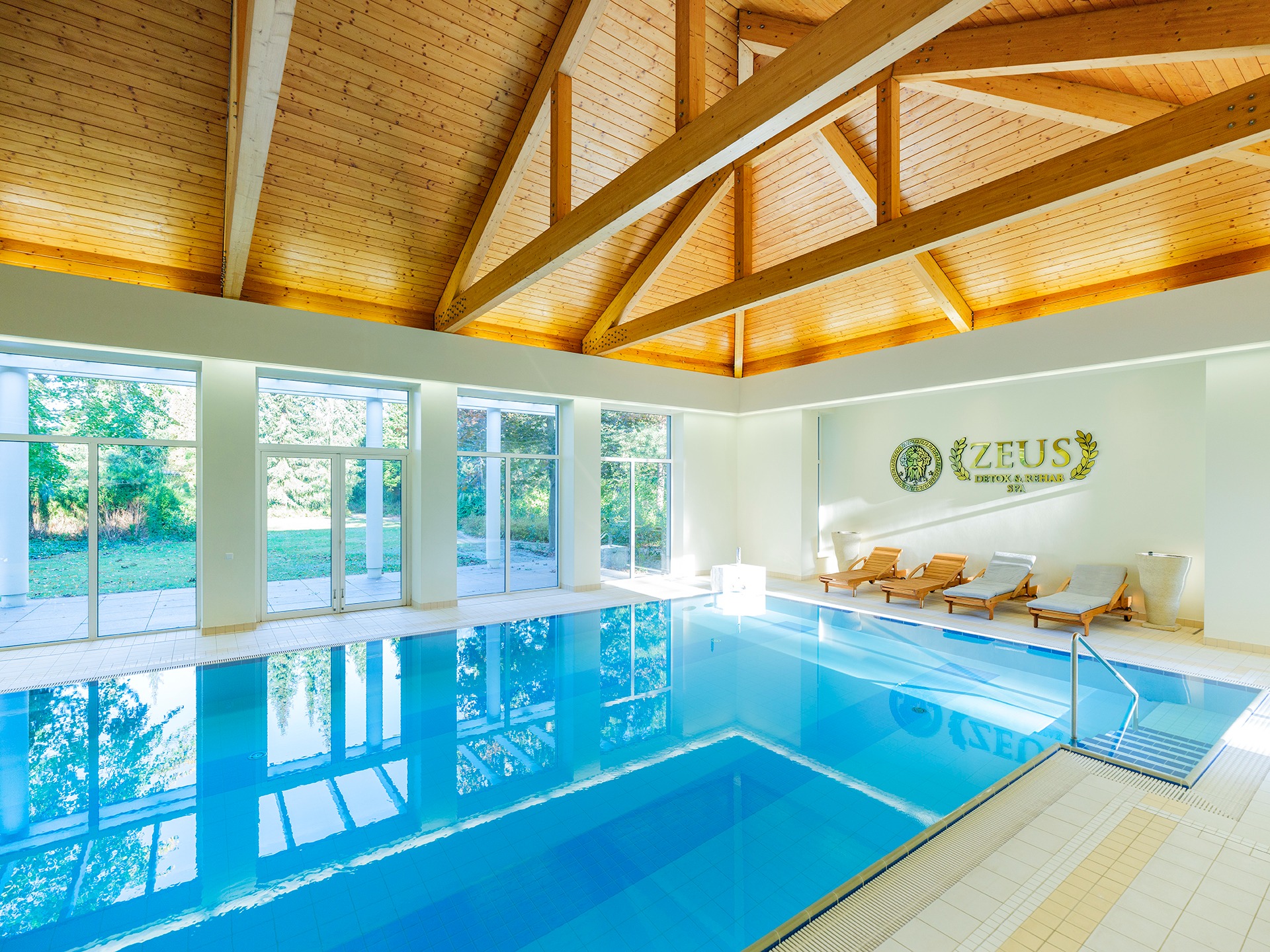 Jasne wnętrze basenu z drewnianym stropem, leżakami i logo 'Zeus Detox & Rehab SPA' na ścianie.