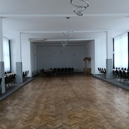 Krzysztof Buczek - Sala z parkietem ułożonym w jodełkę, rzędy krzeseł i stołów po bokach, nowoczesne lampy wiszące. Widok na przestronne, jasne wnętrze.