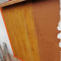 Fragment ściany z częściowo przyklejonym panelem imitującym drewno, widoczna warstwa kleju na pozostałej części ściany, zabezpieczona taśmą malarską.