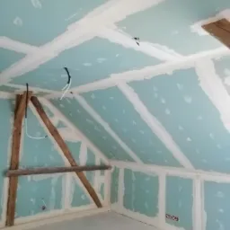 Poddasze w trakcie wykańczania płytami kartonowo-gipsowymi, widoczne drewniane elementy konstrukcyjne, białe szpachlowanie łączeń płyt, wystające przewody elektryczne.