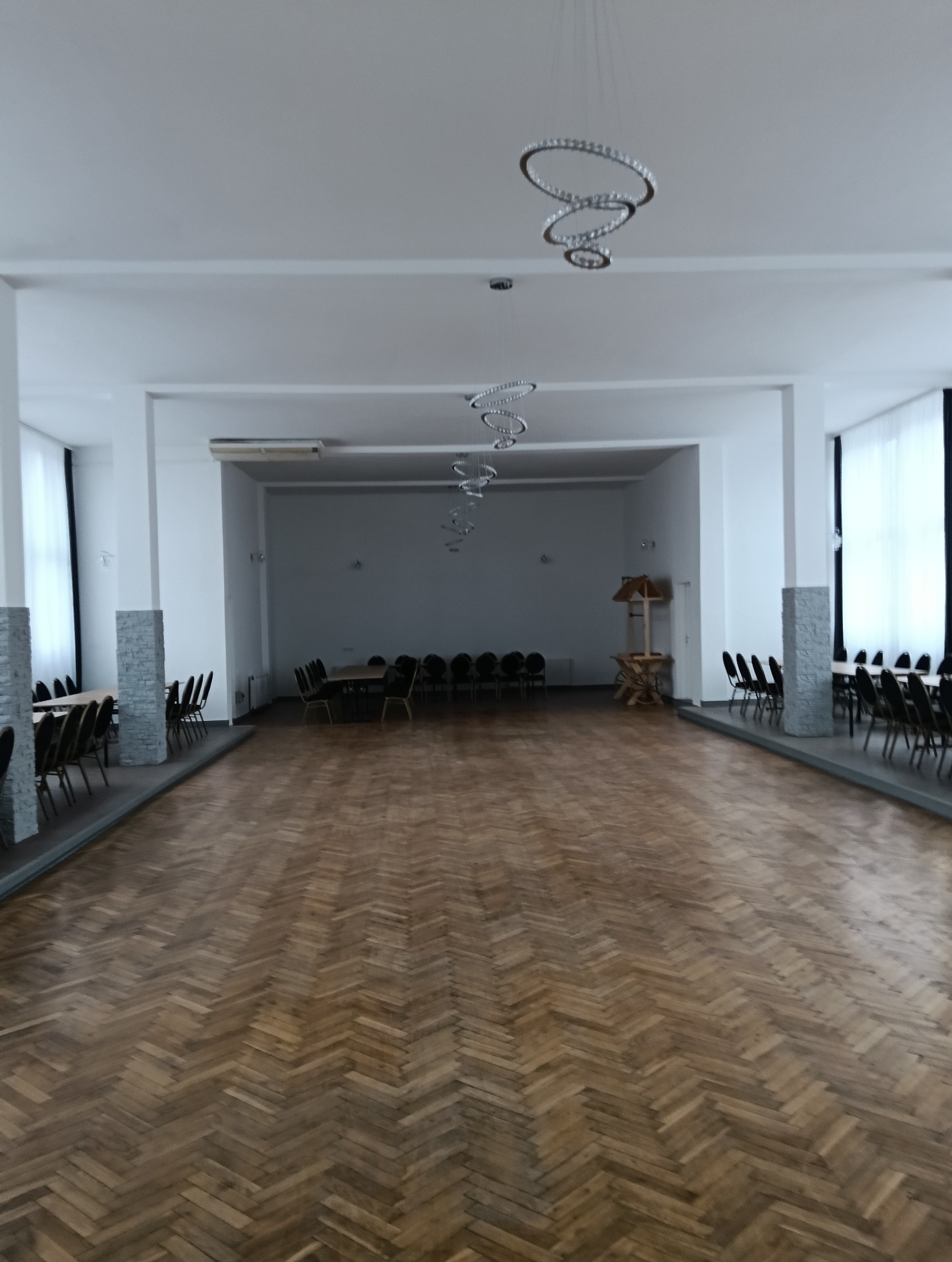 Sala z parkietem ułożonym w jodełkę, rzędy krzeseł i stołów po bokach, nowoczesne lampy wiszące. Widok na przestronne, jasne wnętrze.