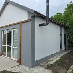 Nowoczesny dom z szarym wykończeniem elewacji, widok z zewnątrz. Elementy drewniane przy oknach, komin zewnętrzny. Ujęcie z niskiego kąta, zieleń w tle.