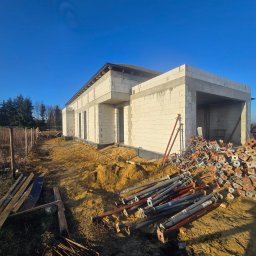 MEGABUD.pl - Nowoczesny dom jednorodzinny z jasnych bloczków w stanie surowym, z otworami okiennymi i garażem, otoczony sprzętem budowlanym pod błękitnym niebem.