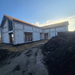 MEGABUD.pl - L-kształtny dom w stanie surowym otwartym, z białymi bloczkami ścian i częściowo zadaszonym dachem, oświetlony promieniami zachodzącego słońca, na tle ziemi budowlanej.