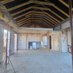MEGABUD.pl - Wnętrze budowanego domu z widoczną drewnianą konstrukcją dachu, białymi ścianami z bloczków i betonowym stropem wspartym na stemplach.