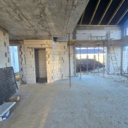 MEGABUD.pl - Wnętrze budowanego domu z widocznymi ścianami z bloczków, betonowym stropem i drewnianą konstrukcją dachu. Na pierwszym planie surowa posadzka i metalowe podpory.