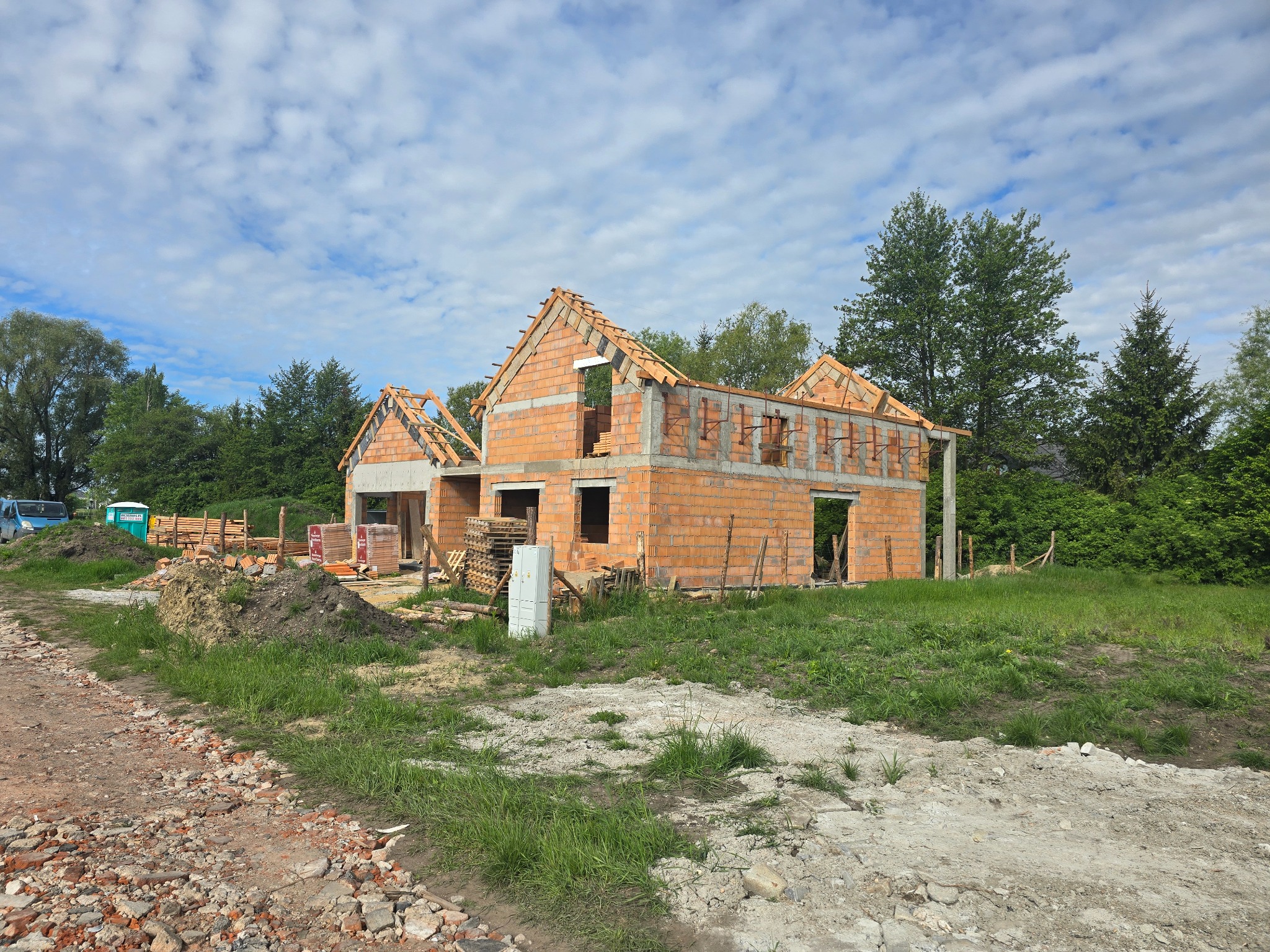 Surowy stan domu z czerwonej cegły w Domasławiu, z widoczną konstrukcją dachu i niezadaszonymi otworami okiennymi, otoczony zielenią i nieutwardzoną drogą.