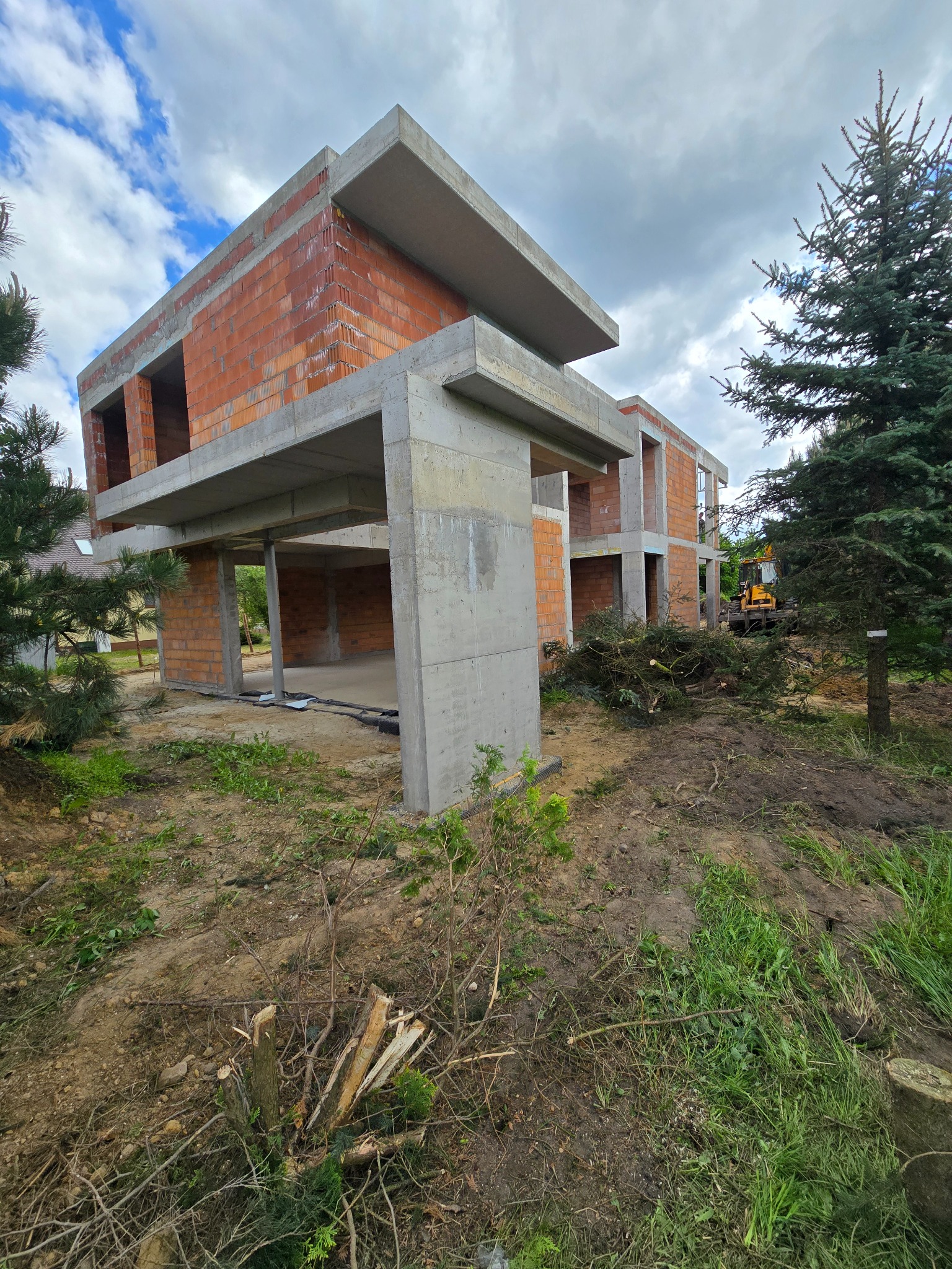 Surowy stan domu z betonu i cegły na działce, widoczne elementy konstrukcyjne, maszyna budowlana w tle, teren z wycinką drzew. Jagodno.