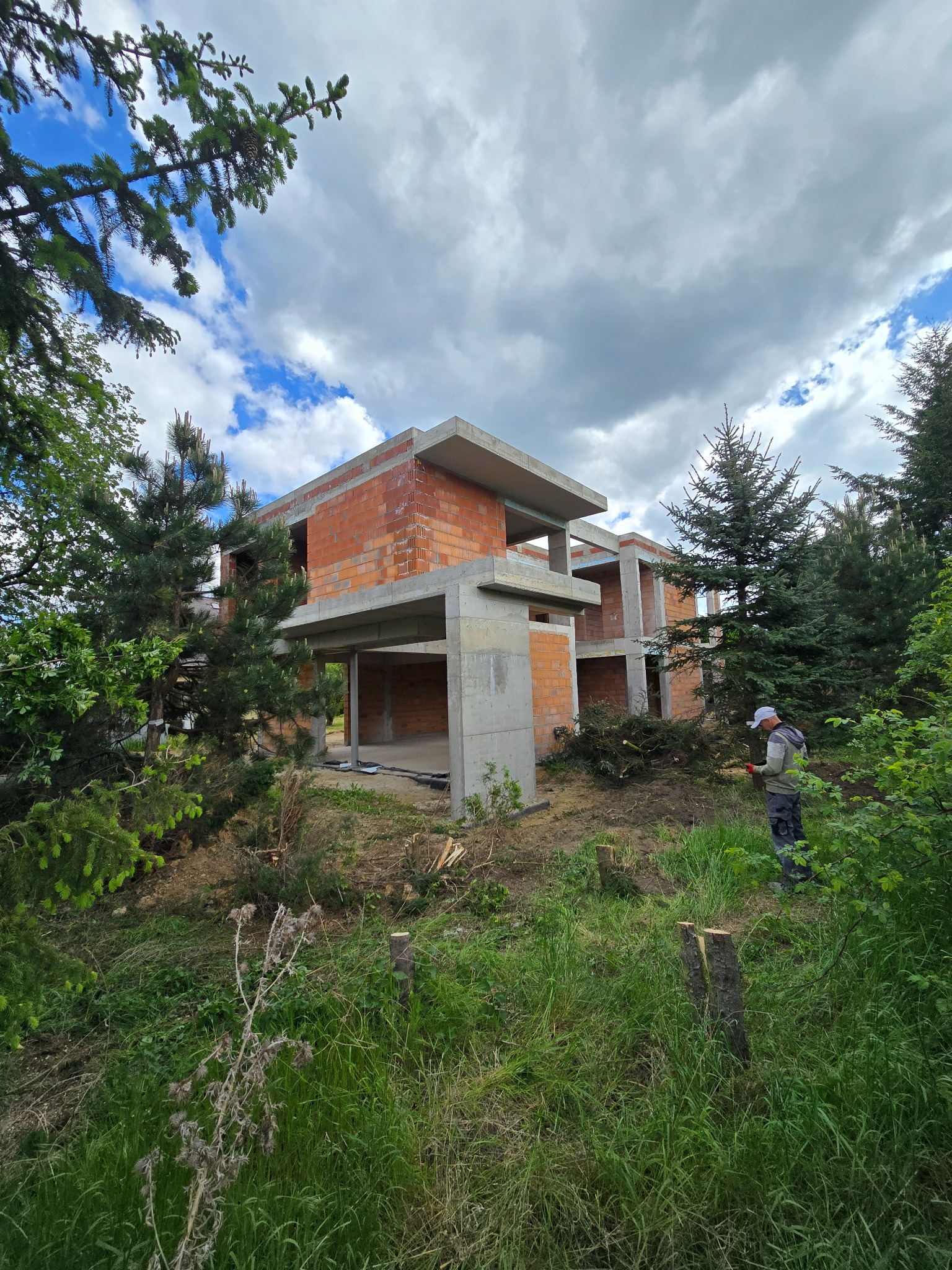 Surowy stan domu z cegły i betonu, otoczonego zielenią. Robotnik w oddali, przygotowuje teren pod dalsze prace budowlane. Pochmurne niebo w tle.