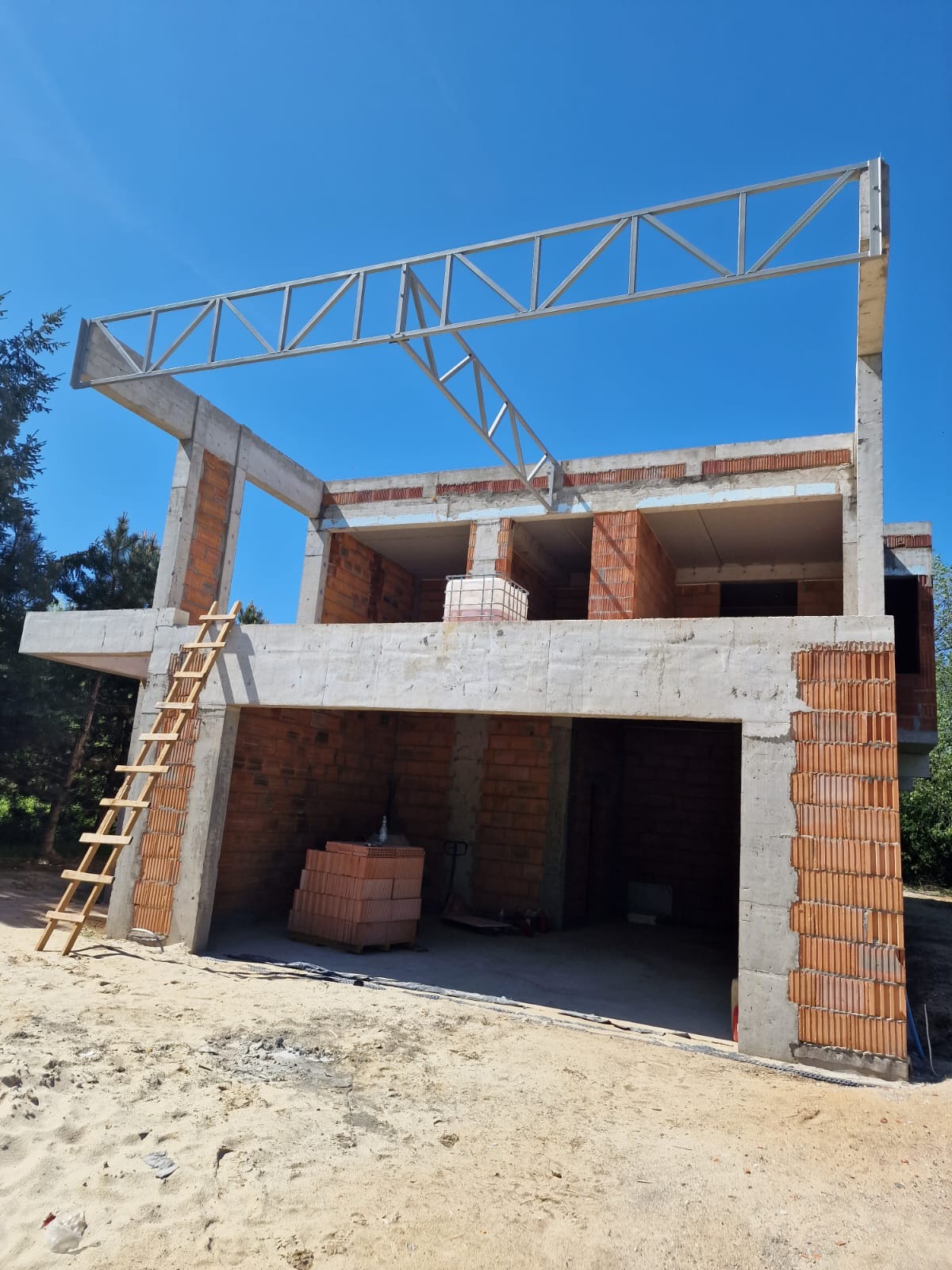 Dwukondygnacyjny dom w stanie surowym otwartym z cegły i betonu, z metalową konstrukcją dachu i drewnianą drabiną opartą o ścianę, na tle błękitnego nieba.