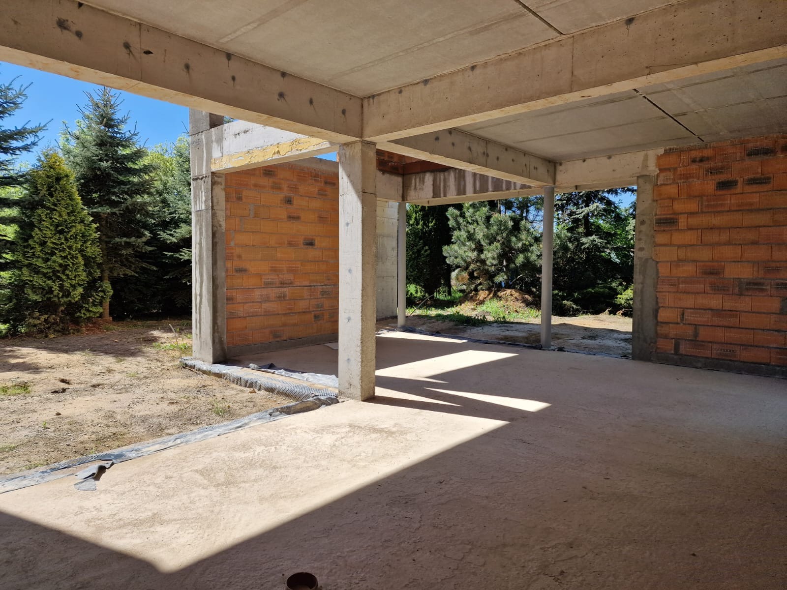 Surowy stan domu z cegły i betonu w otoczeniu zieleni. Widoczne elementy konstrukcyjne i początkowe etapy budowy, z naciskiem na surowość materiałów.