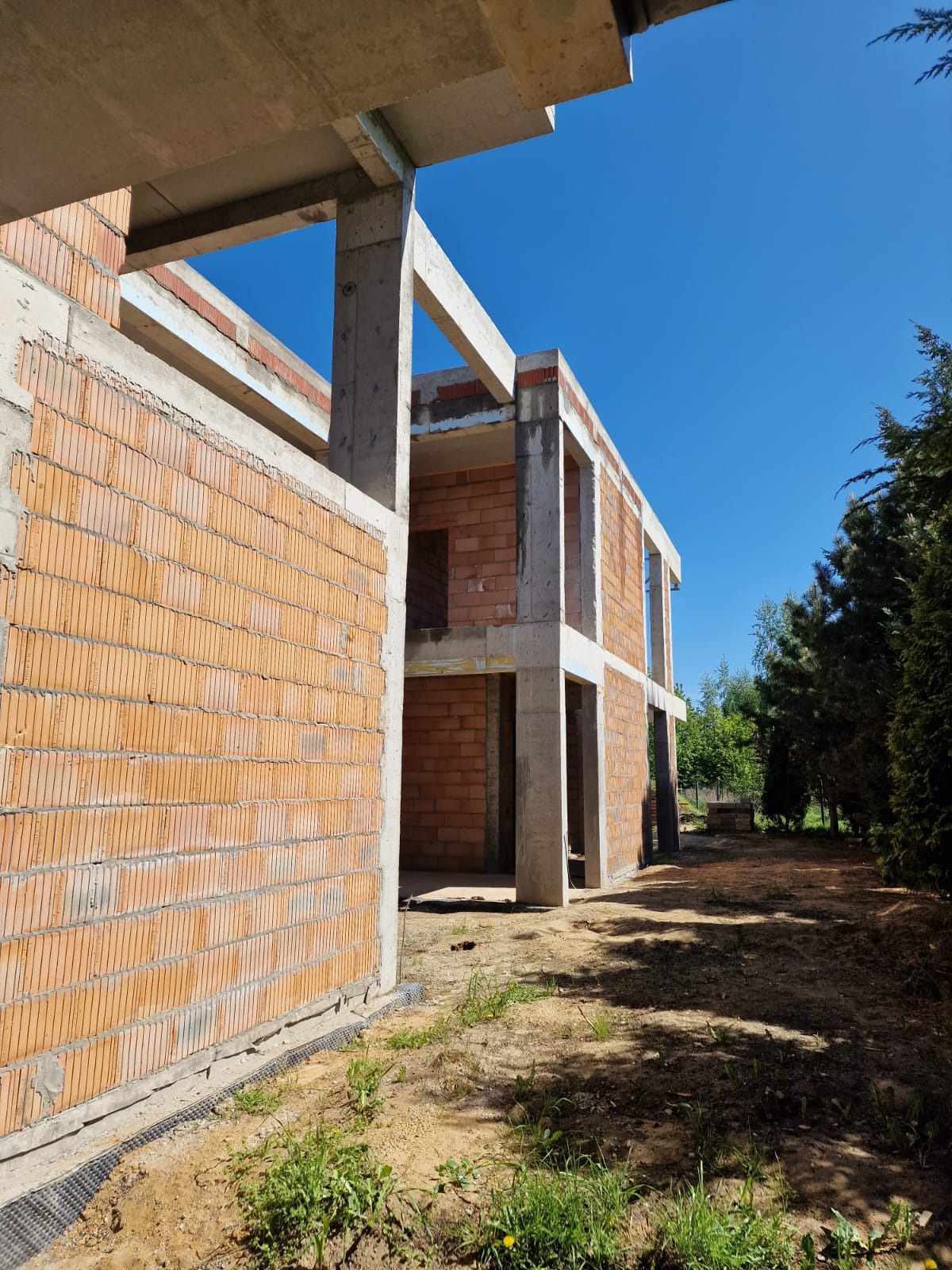 Surowy stan dwukondygnacyjnego domu z czerwonej cegły i betonu w słoneczny dzień. Widoczne zbrojenia i pustaki. W tle zieleń drzew i intensywny błękit nieba.