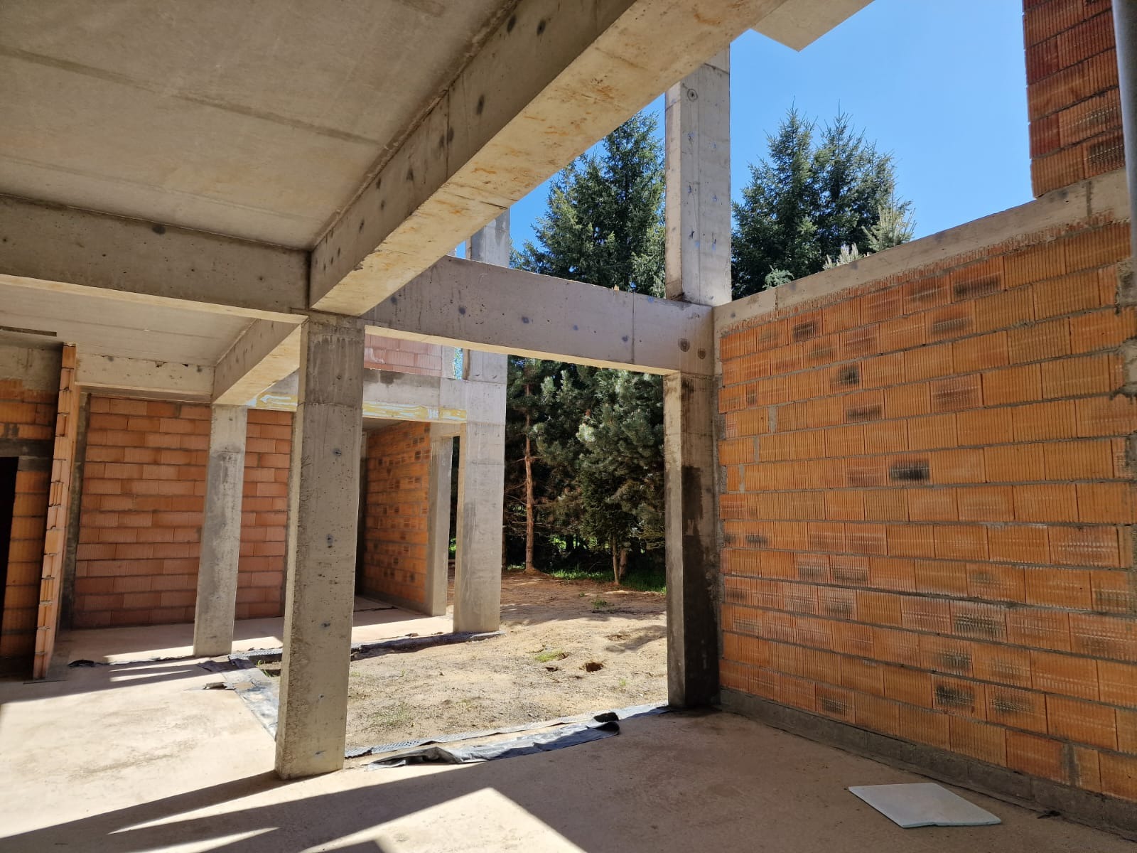 Surowy stan domu w budowie: ceglane ściany, betonowe słupy i belki, widok na zieleń drzew przez otwory okienne. Wnętrze w trakcie realizacji, z widoczną strukturą budynku.