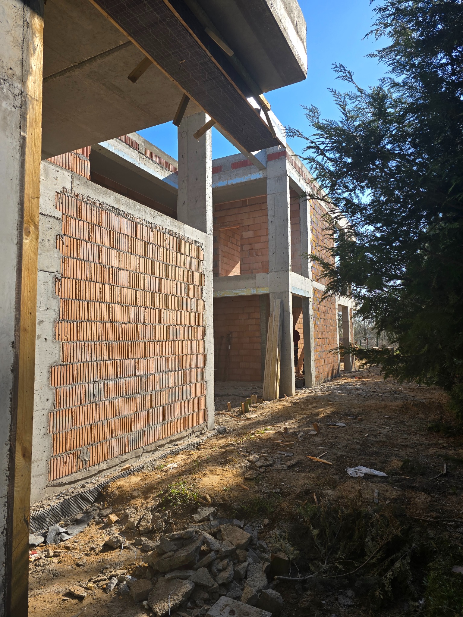 Surowy stan domu w budowie: ściany z czerwonej cegły, betonowe kolumny, widoczne zbrojenia, drzewo iglaste z boku. Niebo w tle.