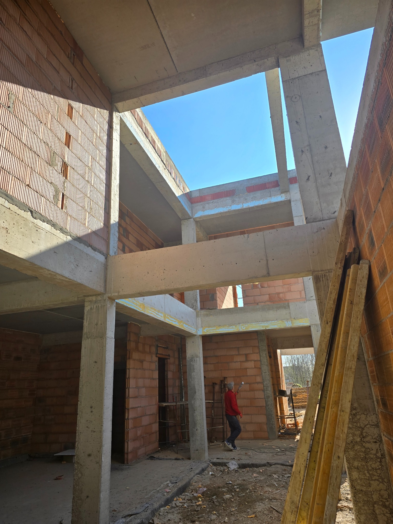 Wnętrze surowego domu z cegły i betonu w trakcie budowy, z widocznym niebem przez otwór w stropie. Osoba w czerwonej bluzie wewnątrz budynku.