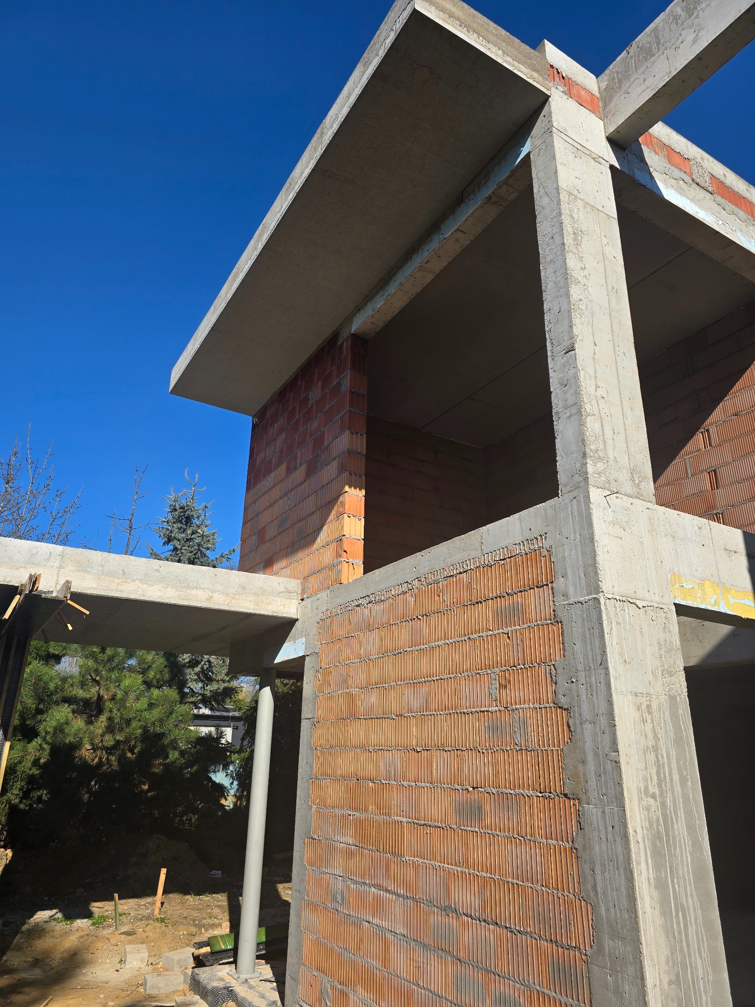 Surowy stan domu z betonu i cegły, widoczne elementy konstrukcyjne, w tle zieleń i błękitne niebo. Kąt ujęcia z dołu podkreśla solidność budynku.