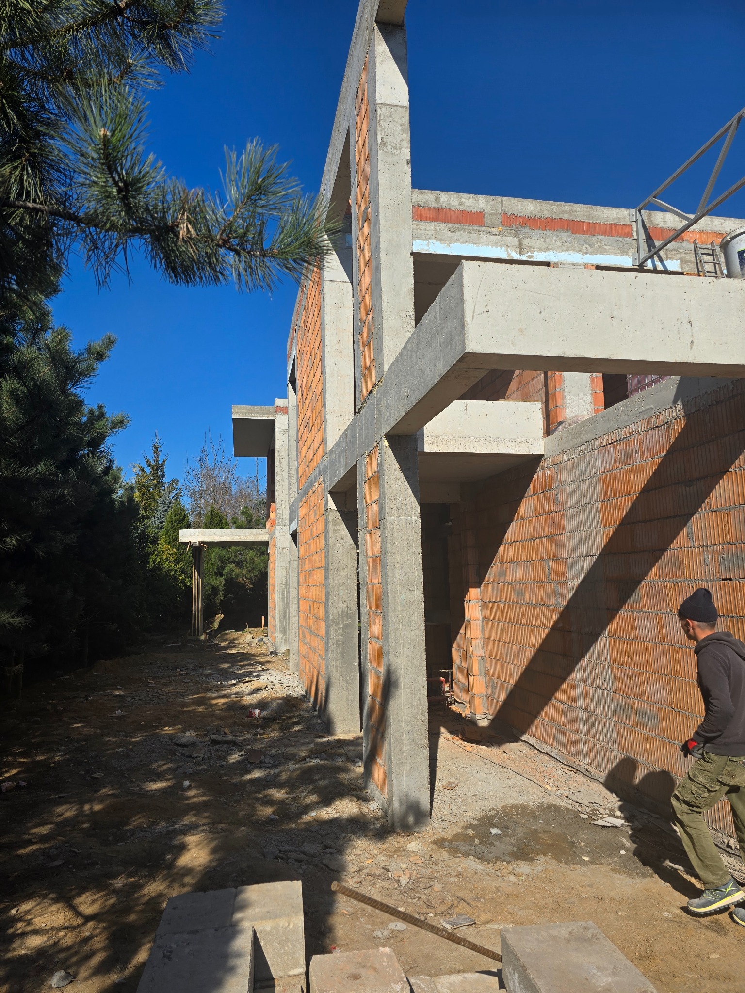 Surowy stan domu murowanego w trakcie budowy: ściany z czerwonej cegły i betonowe słupy konstrukcyjne w jasnym słońcu, pracownik w tle. Widok z zewnątrz.