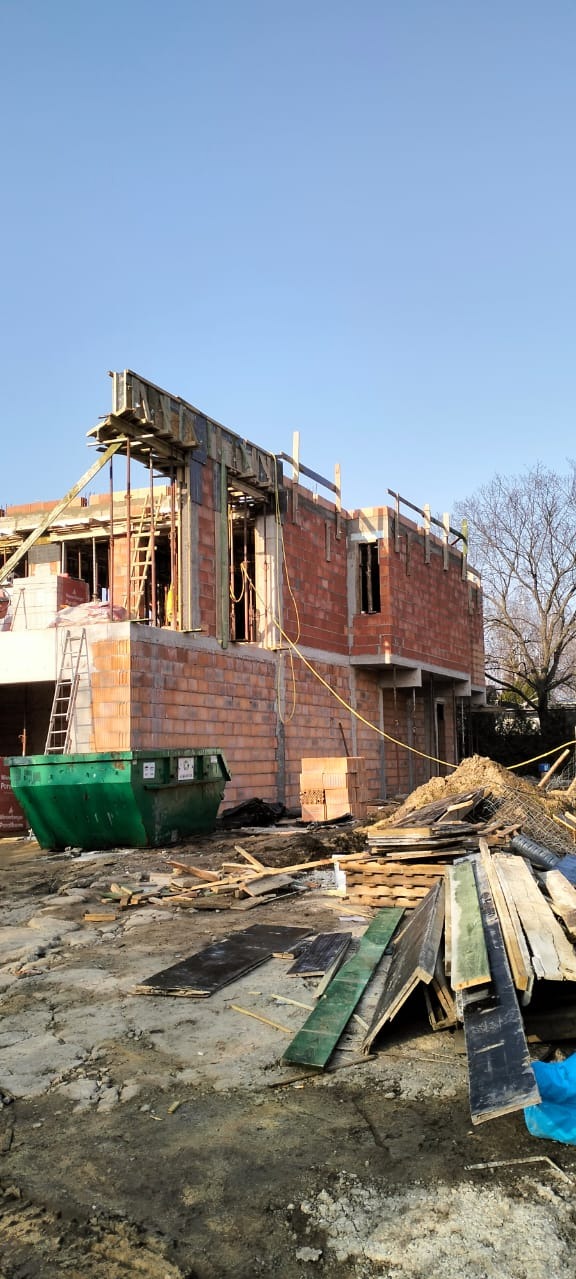 Wznoszenie dwukondygnacyjnego domu z czerwonej cegły. Widoczny kontener na gruz, drewniane deski i szalunki. Budowa w toku, surowy stan.