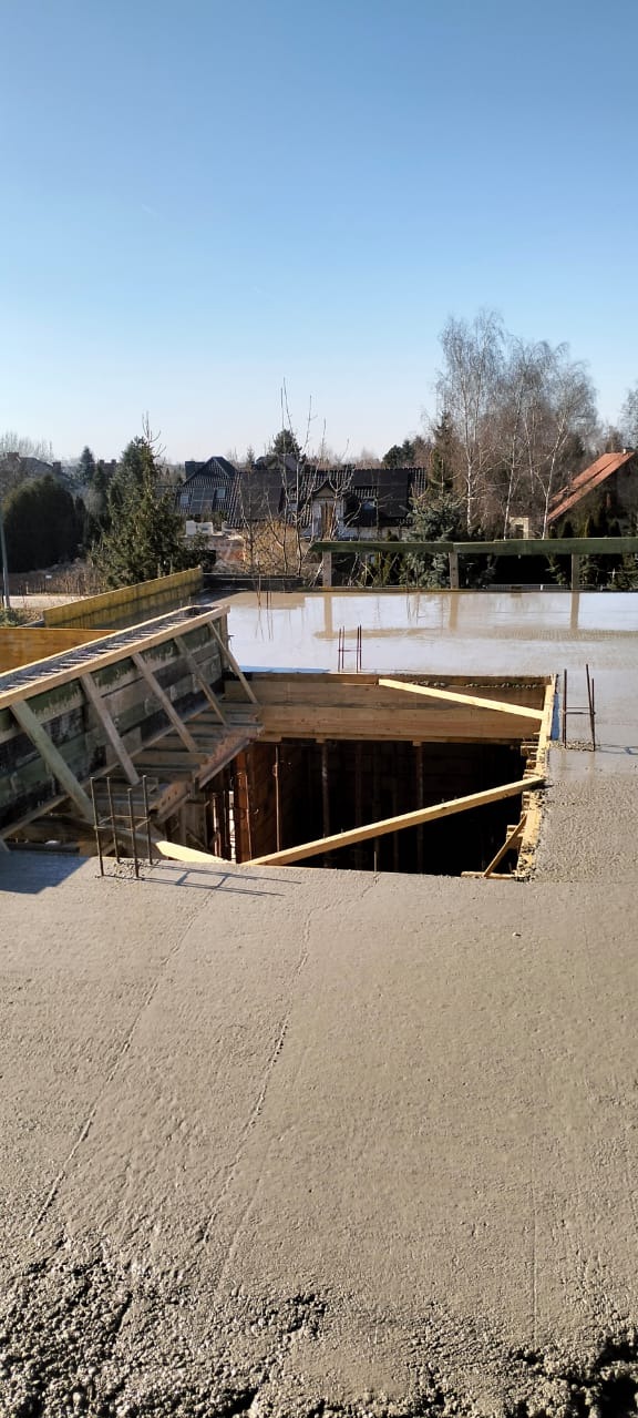 Wylany beton na stropie budynku w trakcie budowy, widoczne drewniane szalunki i wystające pręty zbrojeniowe. W tle zabudowa jednorodzinna i błękitne niebo.