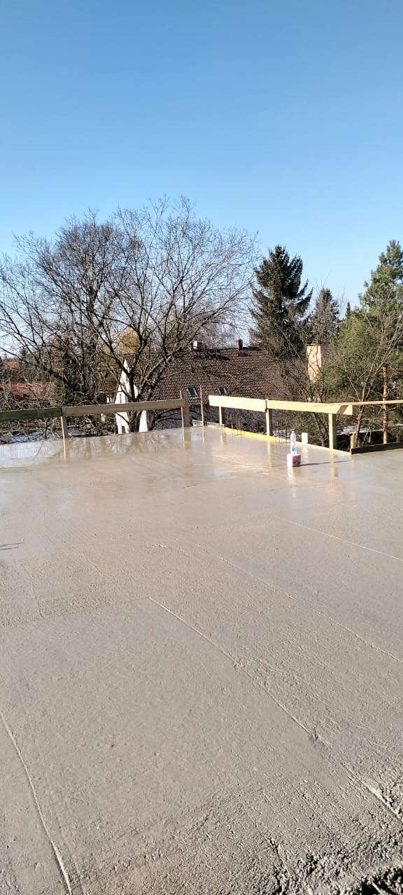 Wylany beton na stropie budynku w trakcie budowy. Widoczne drewniane szalunki i butelka wody. W tle domy i drzewa pod błękitnym niebem.