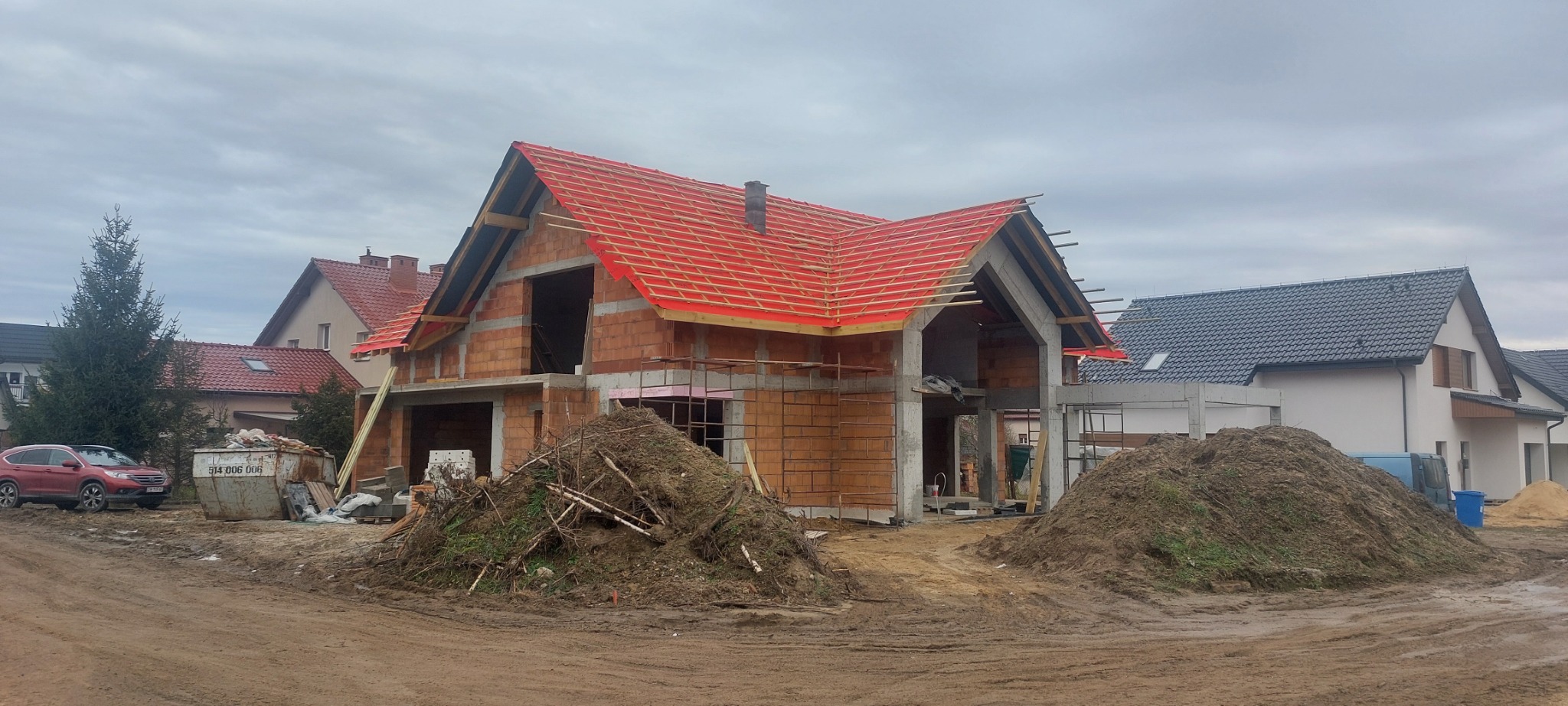Budowa domu jednorodzinnego w trakcie: ściany z cegły, konstrukcja dachu z czerwoną folią, gruz, samochód i kontener na odpady budowlane na pierwszym planie.