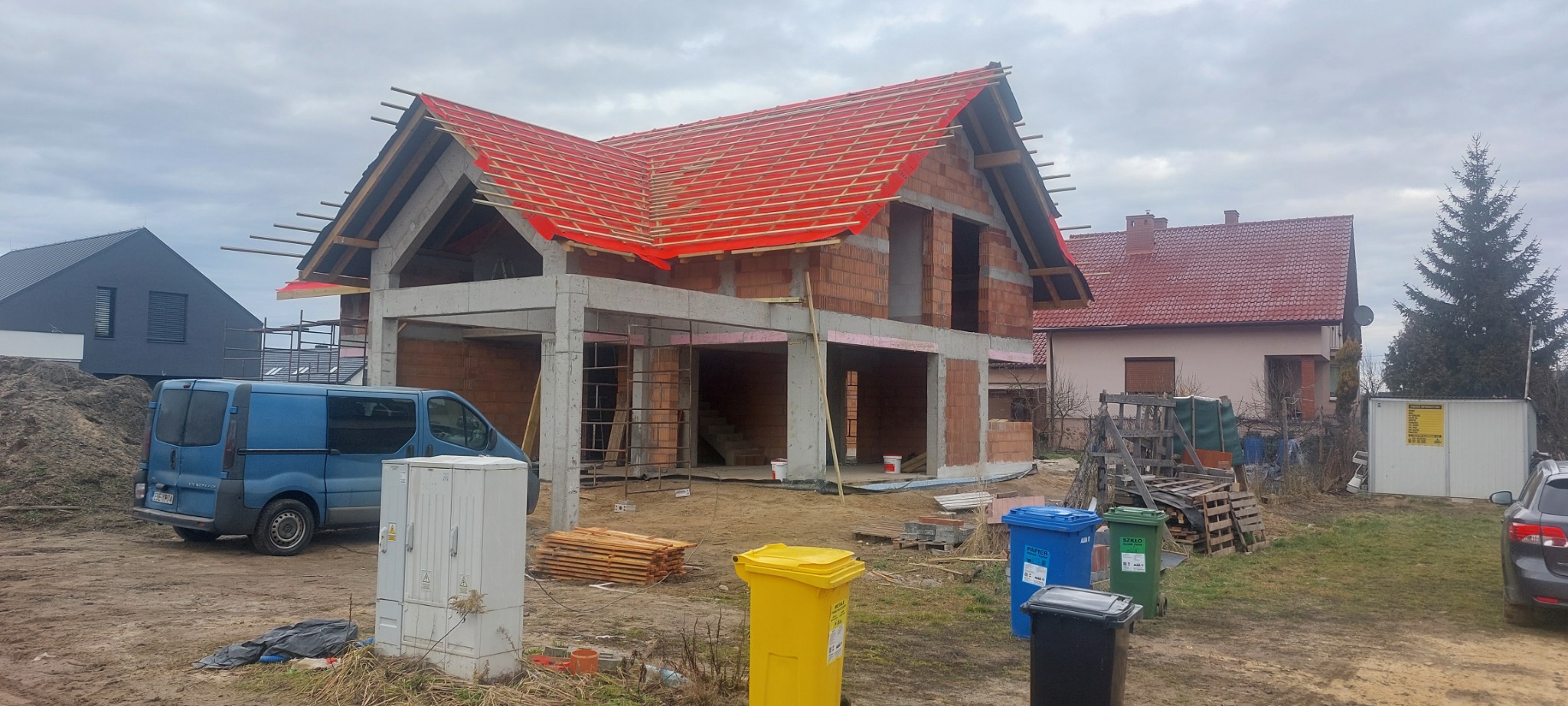 Surowy stan domu z czerwonym pokryciem dachowym w trakcie budowy, z widocznymi pustakami i betonowymi słupami, otoczony gruzem i śmietnikami.