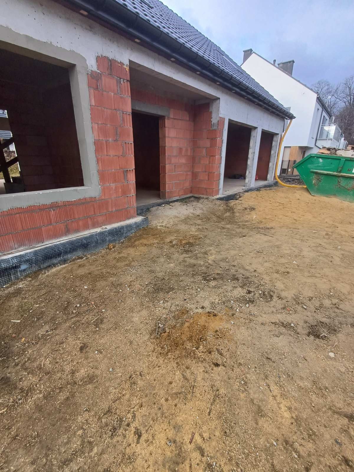 Wznoszenie muru z czerwonej cegły porotherm; widoczne otwory okienne i drzwiowe. Na zewnątrz kontener na gruz i piasek. Budowa domu w Smolcu w trakcie realizacji.