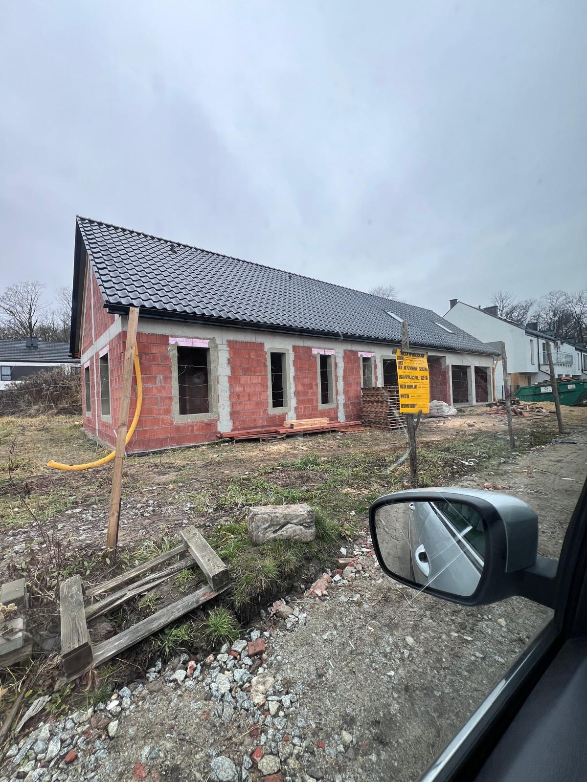 Murowany dom w trakcie budowy z czarnym dachem, widoczna żółta rura i tablica informacyjna. Ujęcie z wnętrza samochodu, widoczne lusterko.