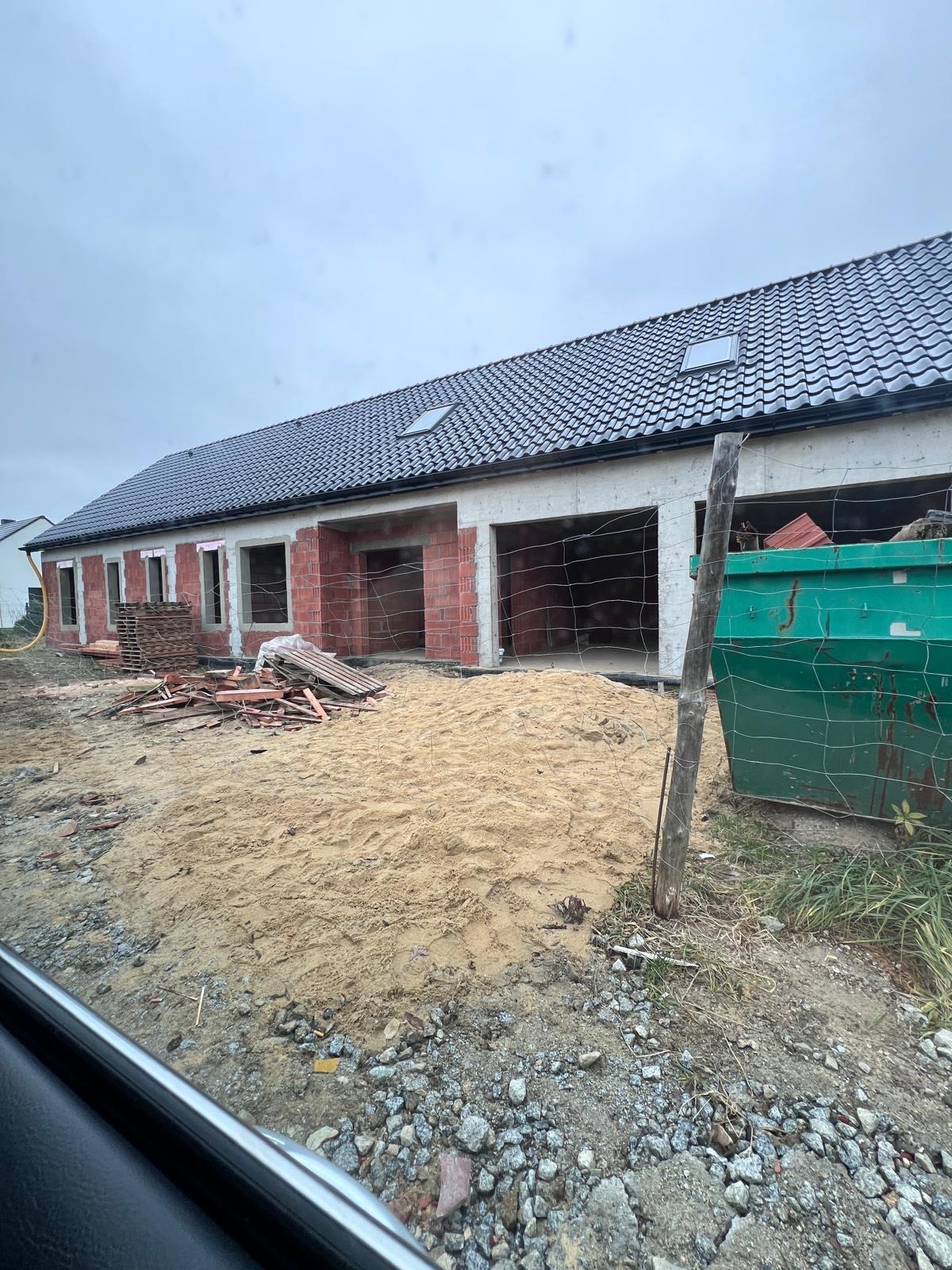 Budowa domu jednorodzinnego w stanie surowym otwartym: widoczne ściany z czerwonej cegły, czarny dach z oknami dachowymi, piasek i kontener na gruz.