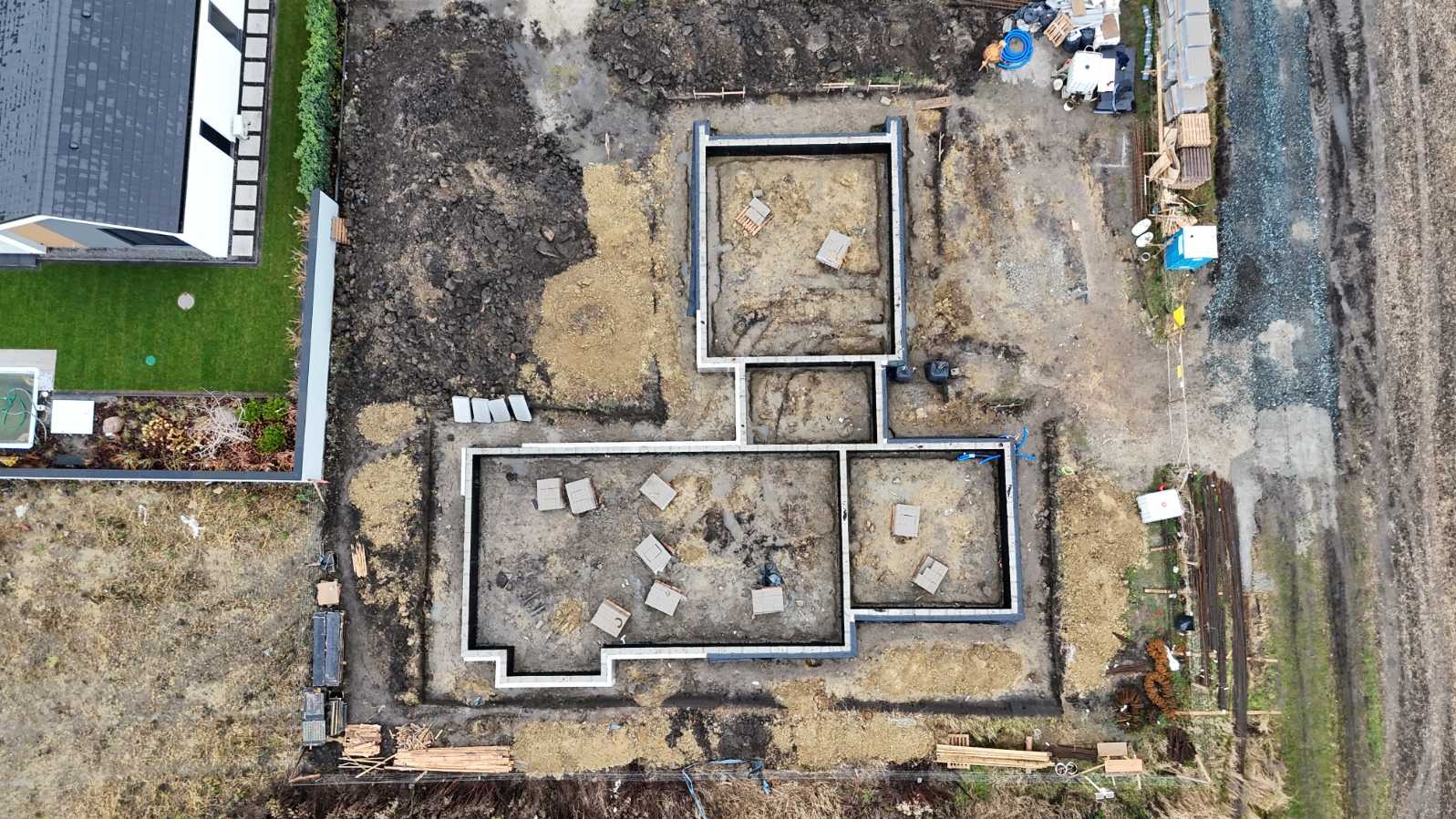 Widok z góry na fundamenty domu jednorodzinnego z zaznaczonymi ścianami, przygotowane do dalszych prac budowlanych, obok widoczny ogród i teren budowy.
