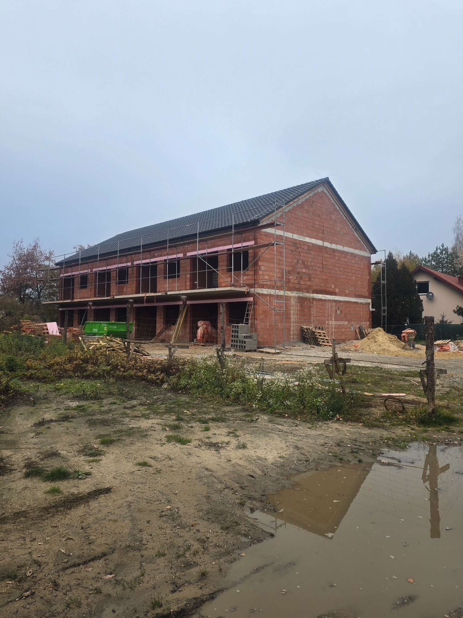 Dwukondygnacyjny dom w trakcie budowy z czerwonej cegły, z częściowo położonym dachem z czarnej dachówki. Widoczne rusztowania i materiały budowlane.