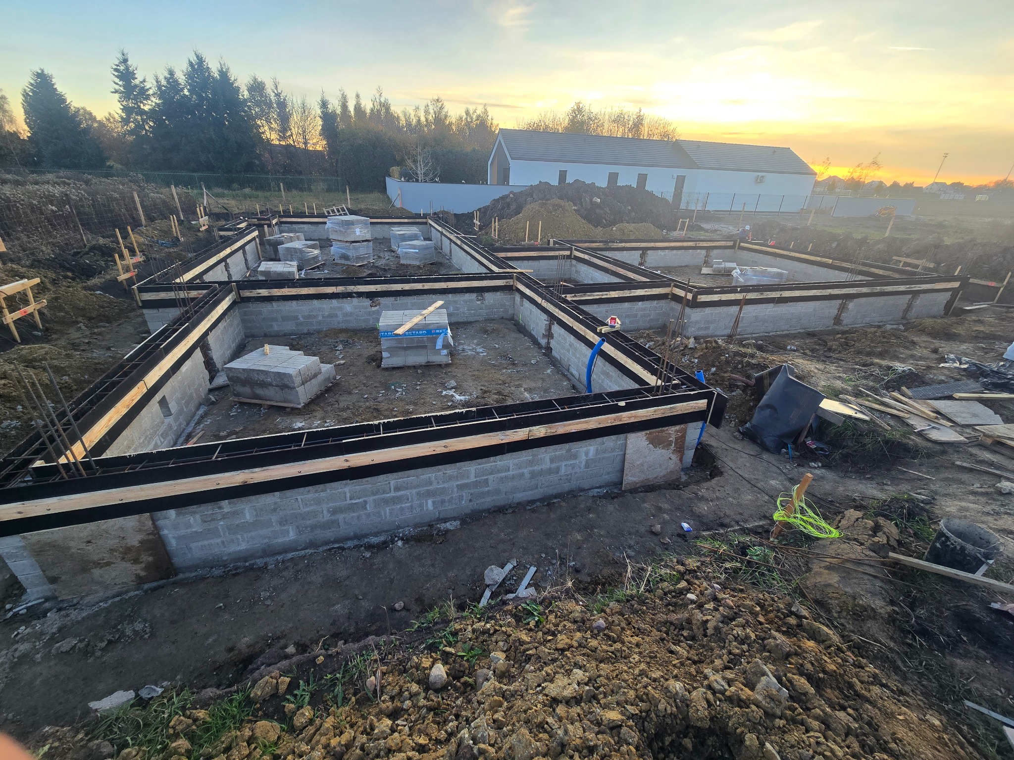 Fundamenty domu z bloczków betonowych, szalunki drewniane, zbrojenie. W tle budynek w trakcie budowy i zachodzące słońce.