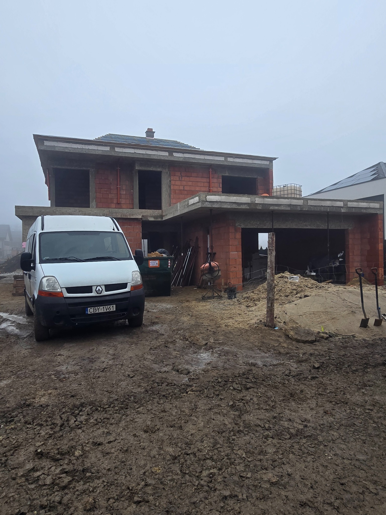 Surowy stan domu murowanego w trakcie budowy, z widocznym szarym niebem i zaparkowanym białym vanem na błotnistym terenie.