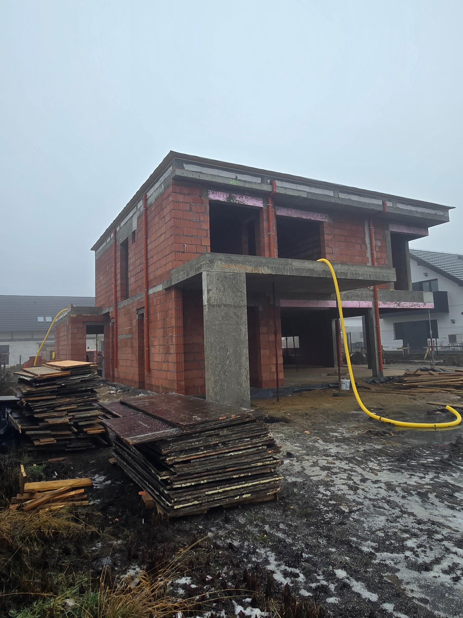 Surowy stan domu murowanego w trakcie budowy, z widocznymi pustakami ceramicznymi, betonowymi nadprożami i stertą płyt szalunkowych na błotnistym terenie.