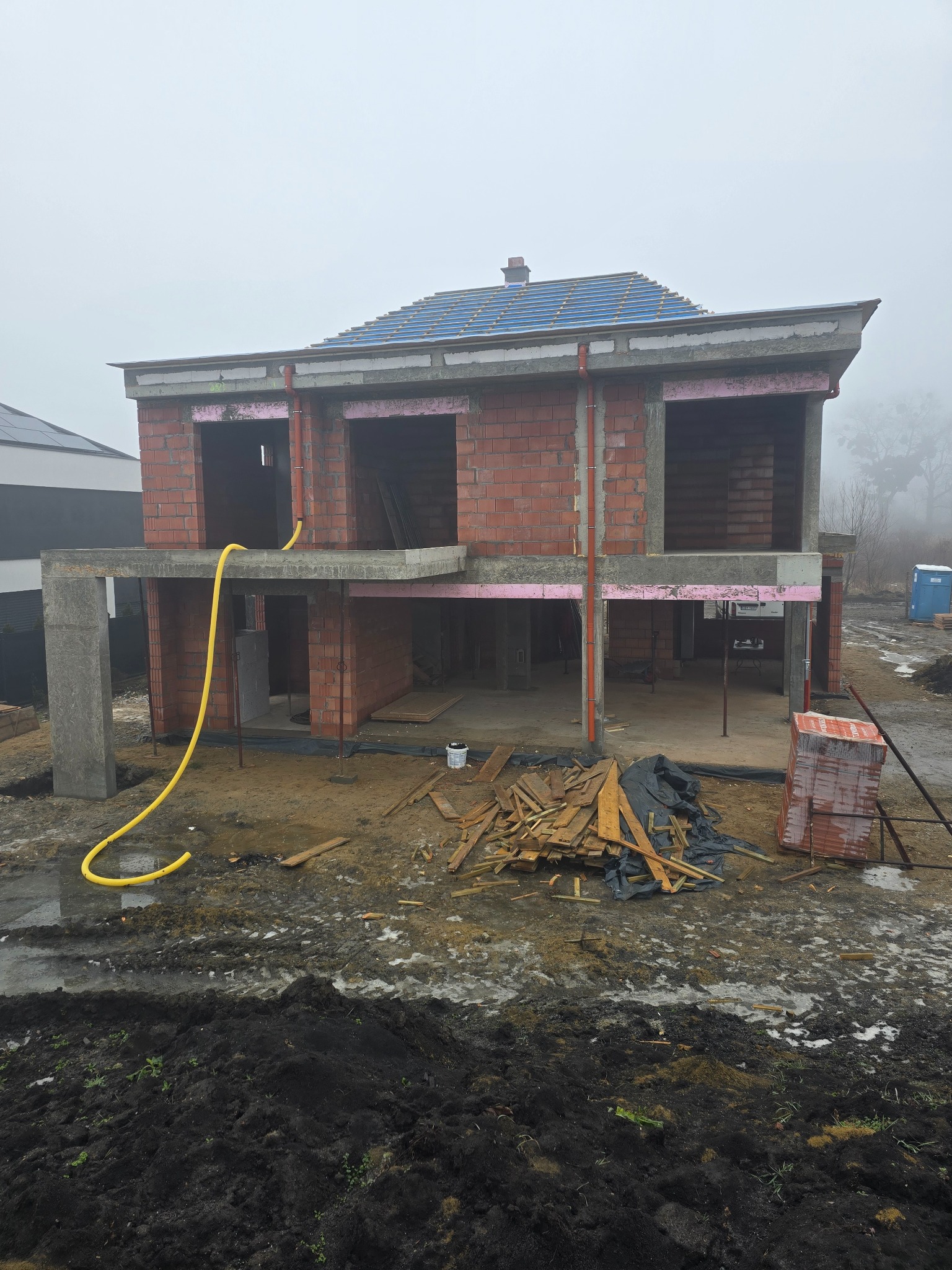 Dwukondygnacyjny dom w stanie surowym otwartym z czerwonej cegły, z częściowo położonym niebieskim dachem, widoczny w mglisty dzień. Na pierwszym planie czarna ziemia.