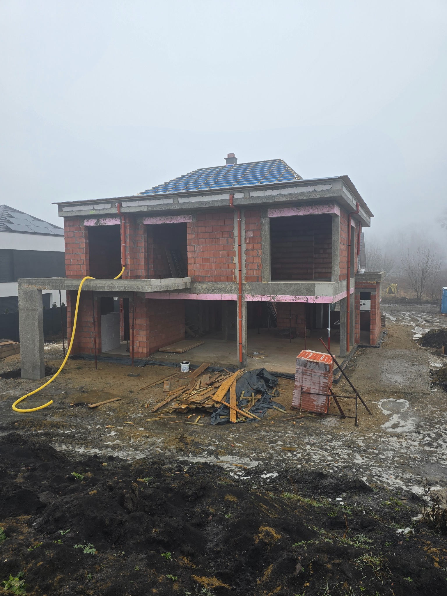 Wznoszenie dwukondygnacyjnego domu z czerwonej cegły w mglisty dzień we Wrocławiu. Widoczne elementy konstrukcyjne i materiały budowlane.