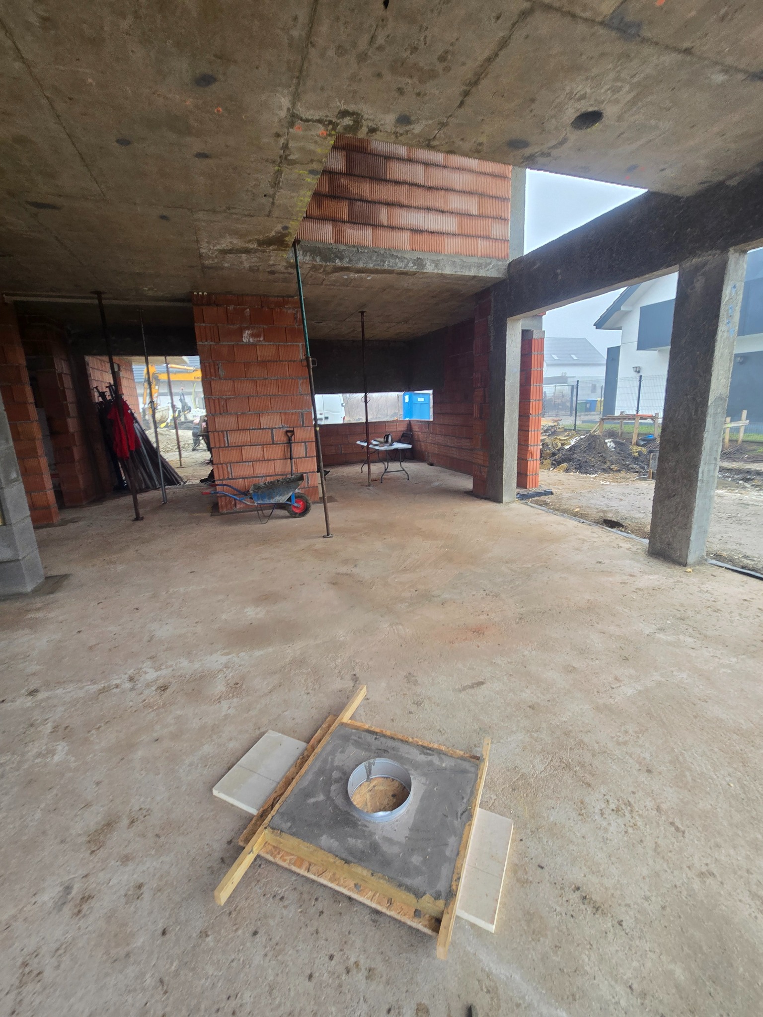 Wnętrze surowego domu w trakcie budowy z czerwonej cegły, widoczne podpory stropu, taczka i narzędzia. Na pierwszym planie element szalunku kominowego.