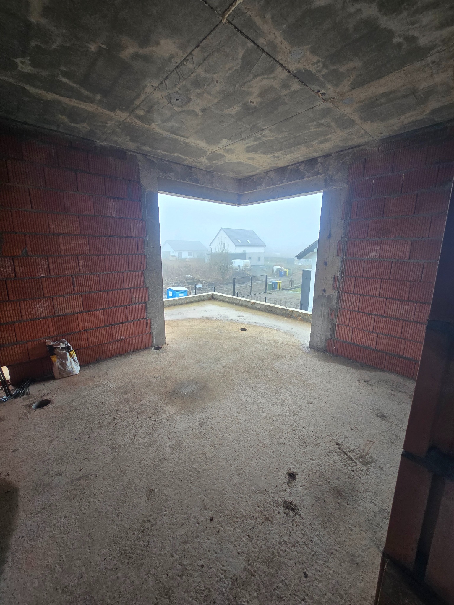 Surowy stan wnętrza domu w trakcie budowy: ściany z czerwonej cegły, betonowa podłoga i sufit, widok na zewnątrz przez otwór okienny w mglisty dzień.