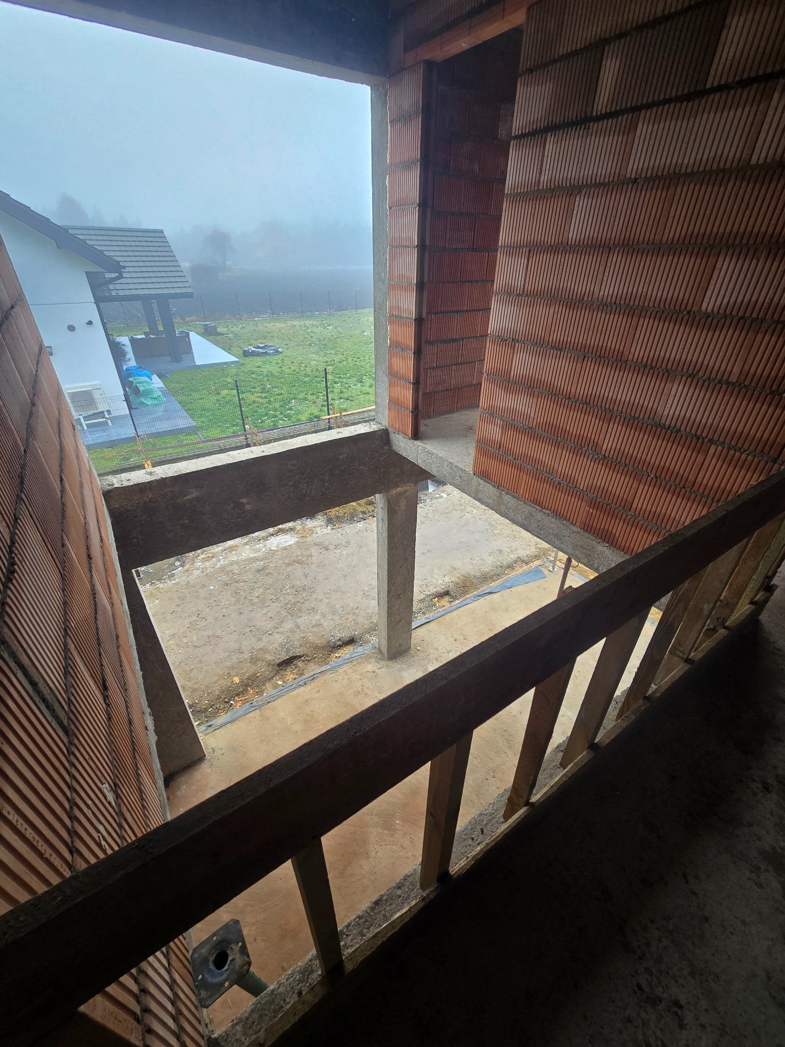 Wnętrze domu w trakcie budowy z cegły ceramicznej, widok z góry na betonowy strop i filar. Na zewnątrz widoczny dom i zieleń w porannej mgle.