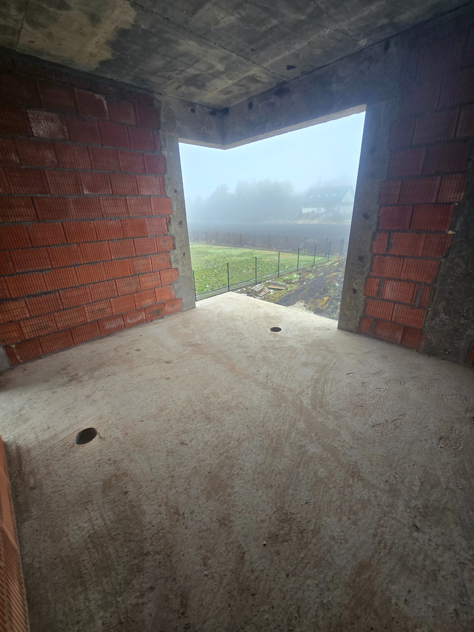 Surowy stan wnętrza domu w trakcie budowy: ściany z czerwonej cegły, betonowa podłoga z otworami, widok na zewnątrz na zewnątrz w mglisty dzień.