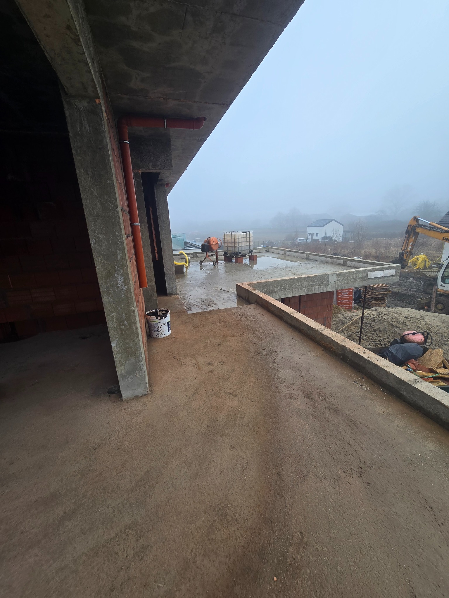 Budowa domu w trakcie realizacji: wylany fundament, widoczny beton, cegły, rury, mieszalnik i koparka w tle, mglista pogoda.