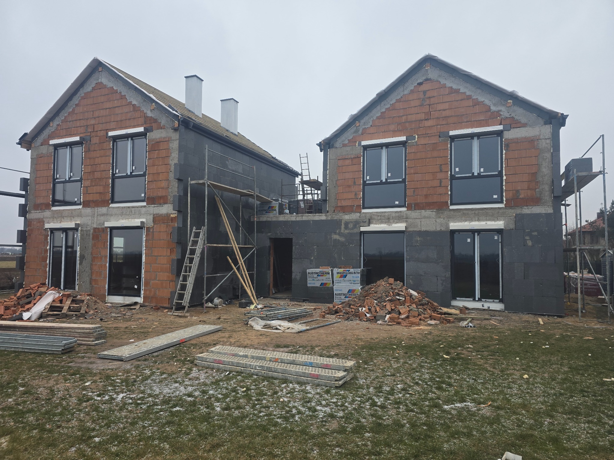 Budowa bliźniaka: surowy mur z oknami, ocieplenie grafitowym styropianem, rusztowania. Cegły i materiały budowlane na trawie. Etap prac przed elewacją.